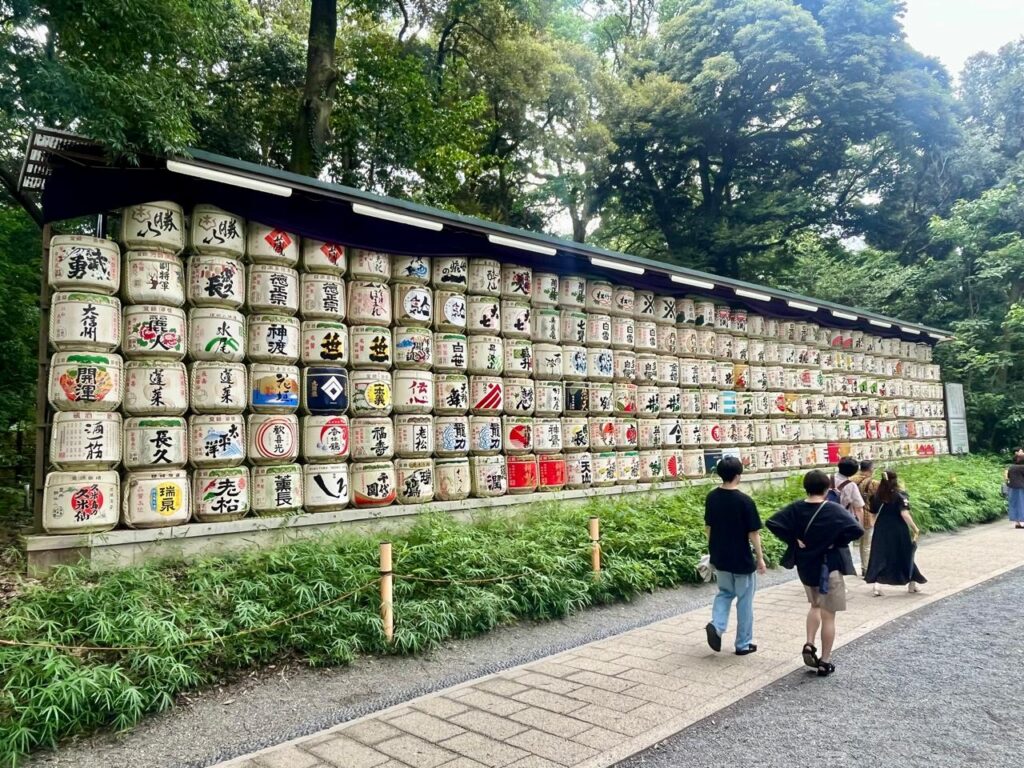 Barriles de sake decorativos alineados junto al sendero del santuario Meiji Jingu en Tokio, rodeados de vegetación y visitantes caminando.